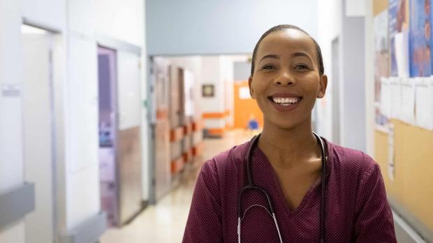 A nurse in red scrubs and a stethoscope stands in a hospital corridor and smiles at the camera