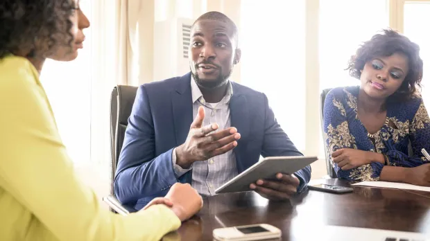 Professional Indemnity - business man gives advice A professional man in a tailored suit sits at a table with two business women. He is holding a tablet computer and speaks directly to one of the women.