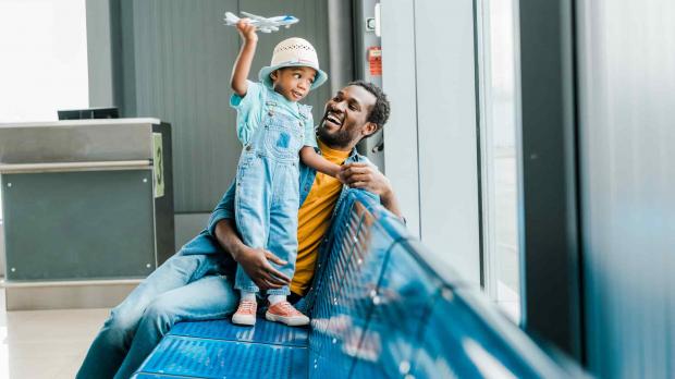 A dad and his little son sit on a bench at the airport terminal. The little boy stands on the bench and holds a toy plane above his head