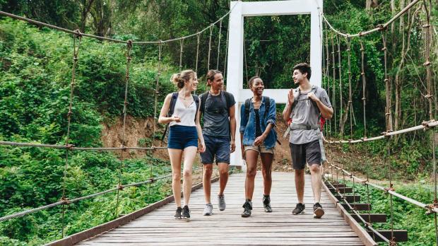 Four young professionals walking across a suspension bridge in a lush green forest, wearing backpacks and talking.