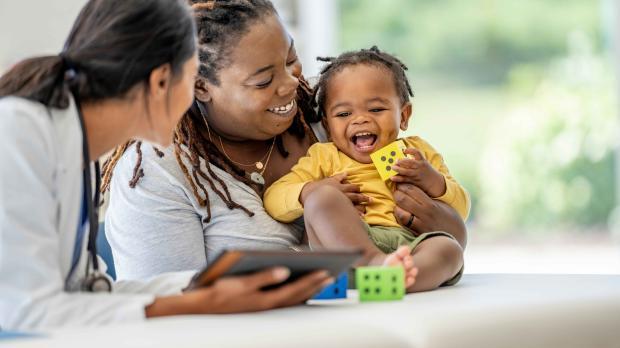 Mother holding a smiling toddler while speaking with a healthcare professional during a clinic visit.
