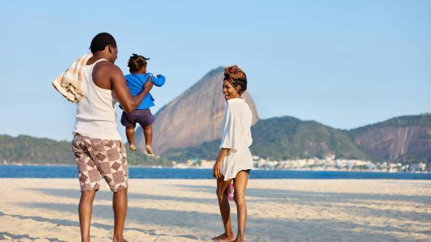 Young family enjoying time together on a sunny beach with ocean and mountains in the background.