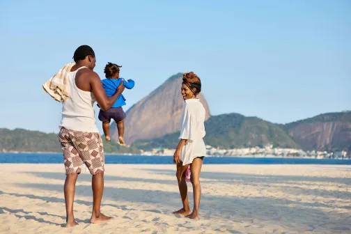 Young family enjoying time together on a sunny beach with ocean and mountains in the background.