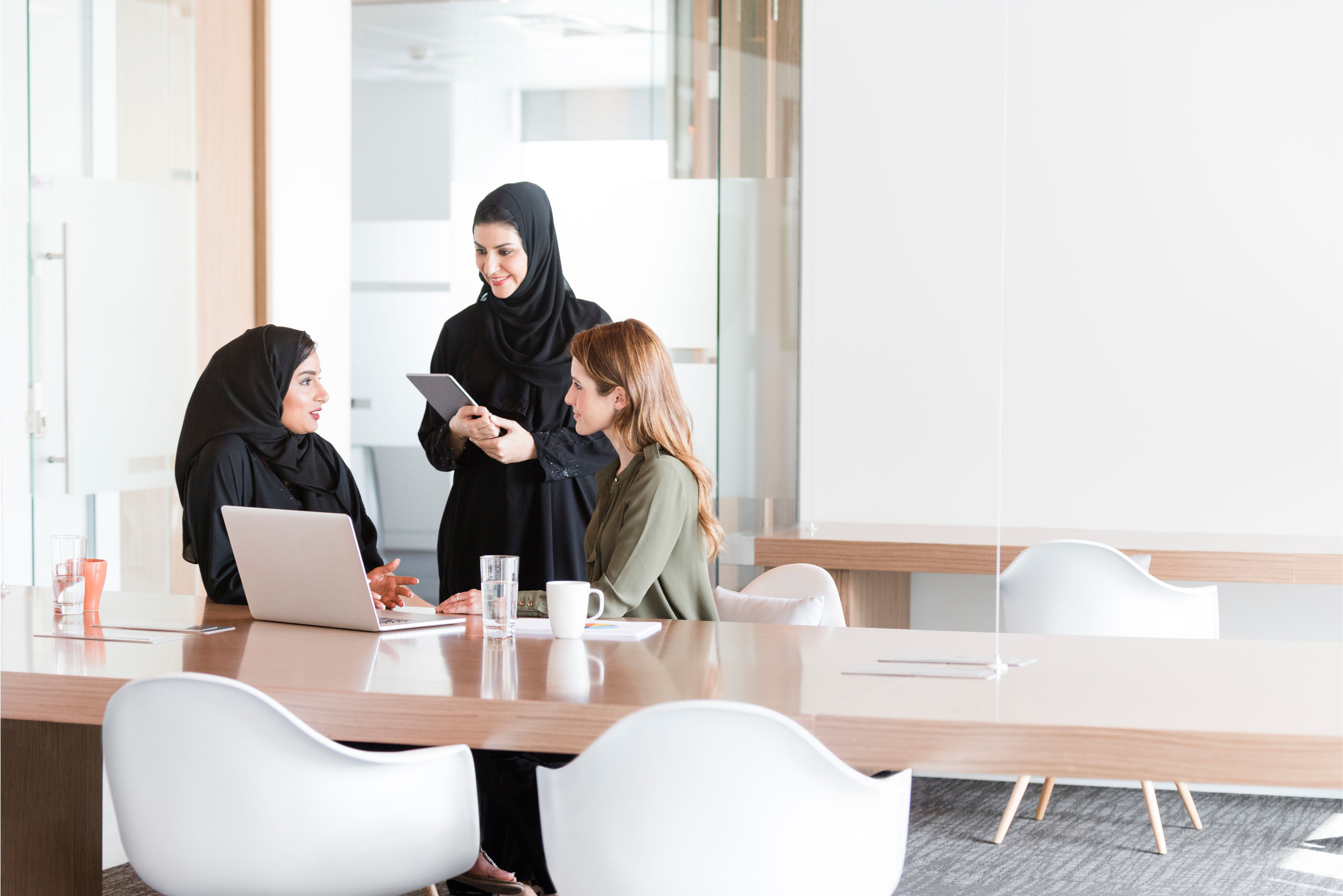 Three female professionals, including Emirati women in abayas, discussing employee benefits in a bright modern office — representing collaboration and engagement in the UAE workplace.
