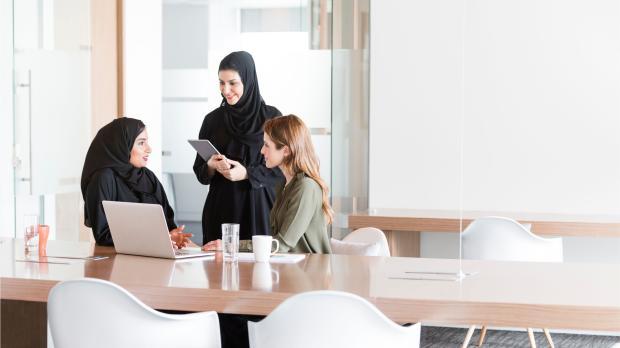 Three female professionals, including Emirati women in abayas, discussing employee benefits in a bright modern office — representing collaboration and engagement in the UAE workplace.