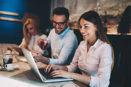 Smiling colleagues working together on a laptop in a relaxed café-style setting, symbolising teamwork, employee engagement, and a positive workplace culture.