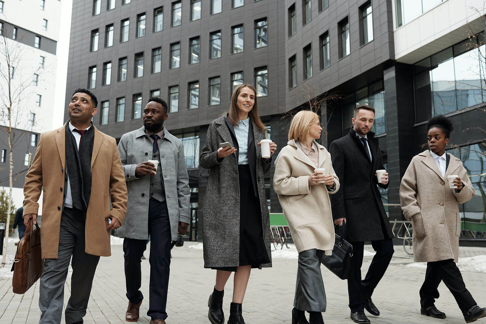 Six business professionals in winter coats walk together through a modern office complex courtyard, holding coffee cups and briefcases, chatting and smiling against the backdrop of tall, dark-glass buildings and patches of lingering snow.