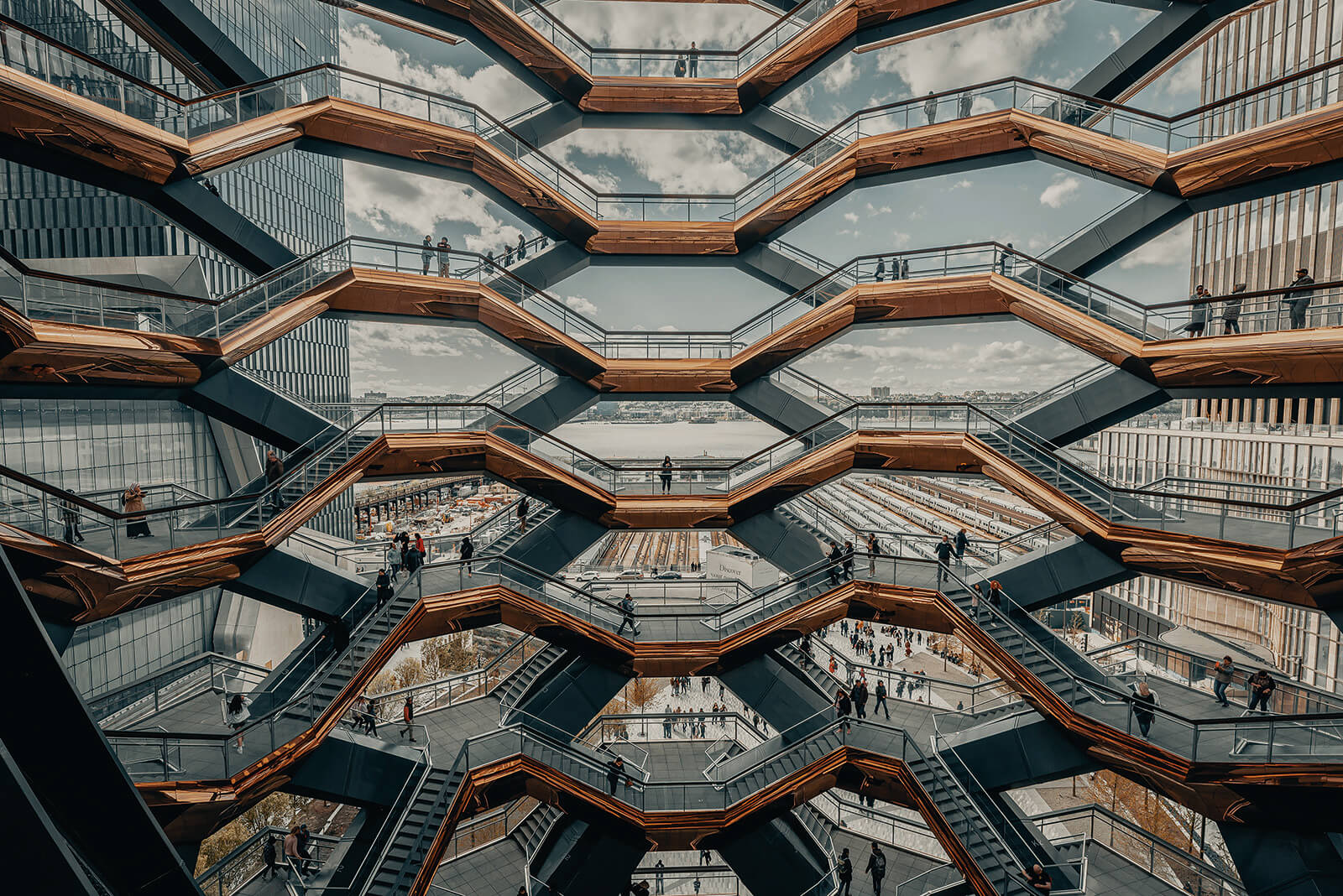 Interior view of a honeycomb-like multi-level staircase structure with zigzagging walkways, where small groups of people walk and stand, framed by modern glass buildings and a partly cloudy sky in the background.