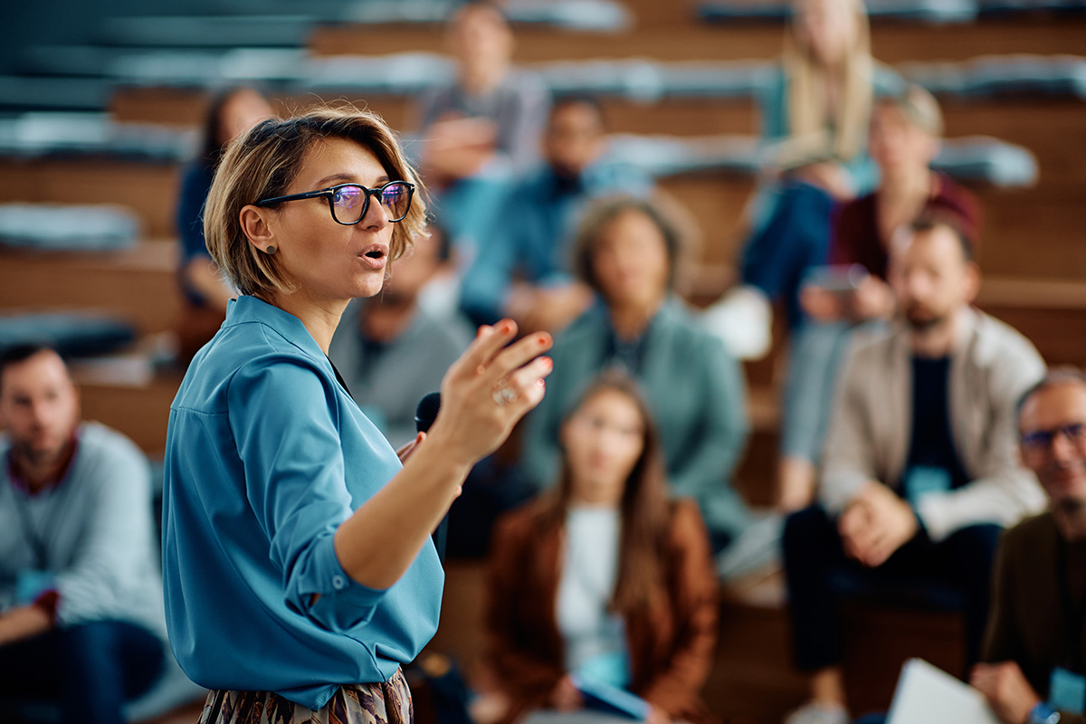 A person in a blue blouse and glasses stands speaking in front of a blurred audience seated in tiered rows, gesturing with one hand while holding a microphone.