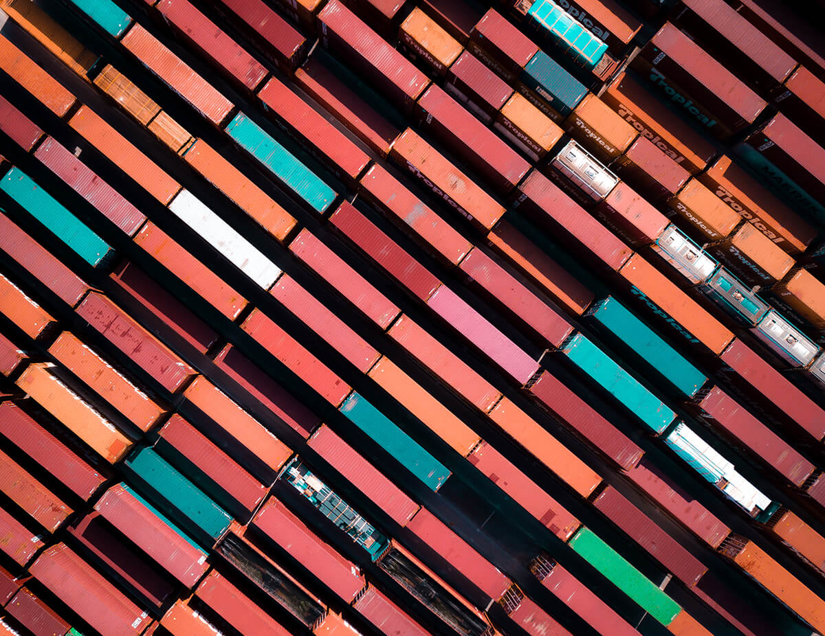 An aerial, diagonal view of tightly packed shipping containers in various colors—red, orange, teal, and white—stacked in parallel rows, creating a bold geometric pattern across the frame.