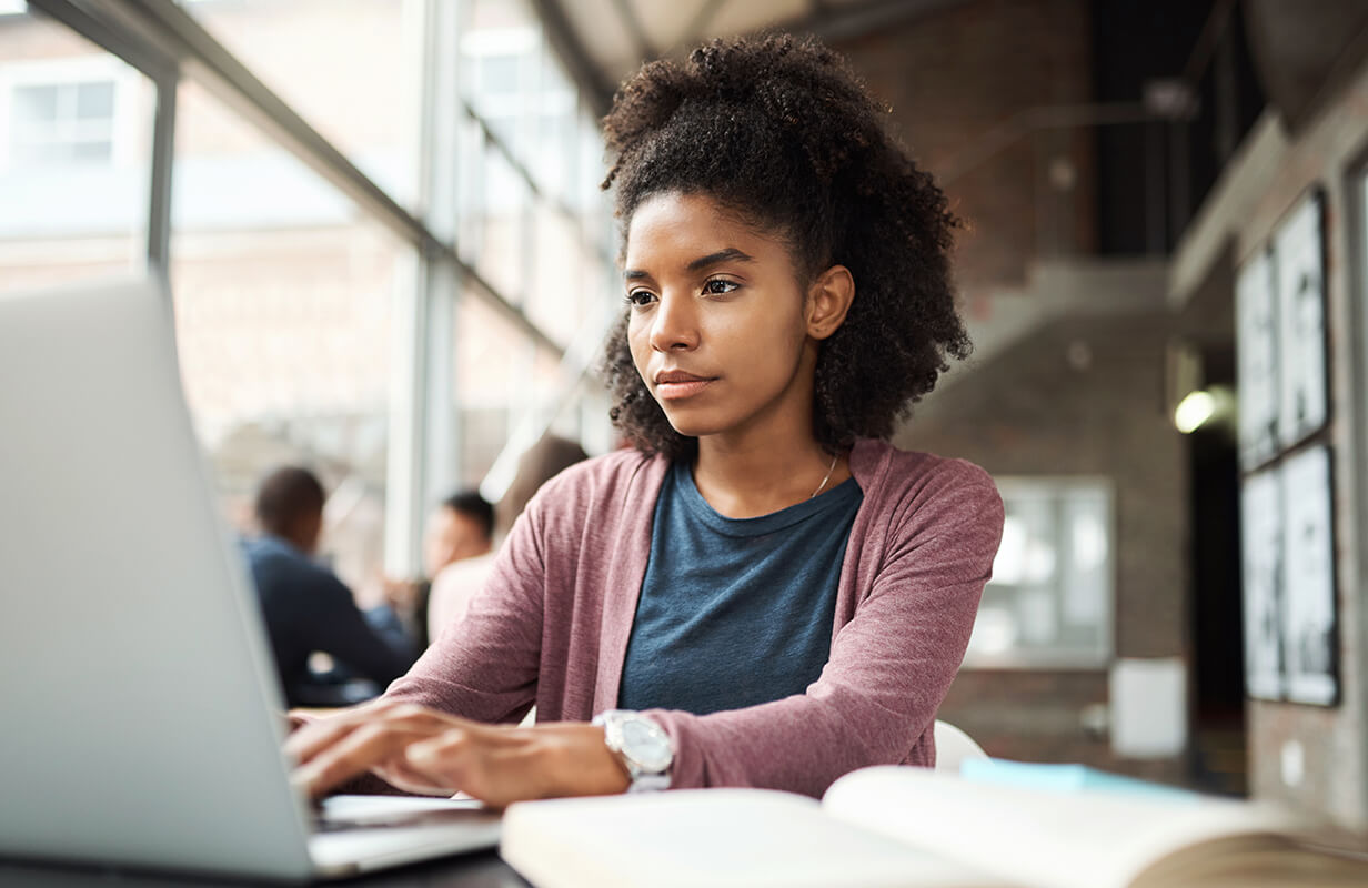 A young woman works intently on a laptop in a bright, modern open office space, with a large window beside her and an open book in the foreground.
