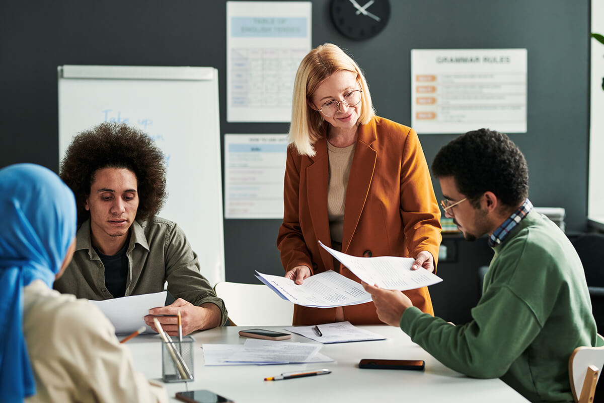 An adult education classroom scene where a middle‑aged instructor in a rust-colored blazer stands at a table, handing out worksheets to a group of diverse adult students seated around her, with notebooks and phones on the table and a “GRAMMAR RULES” poster and whiteboard visible in the background.