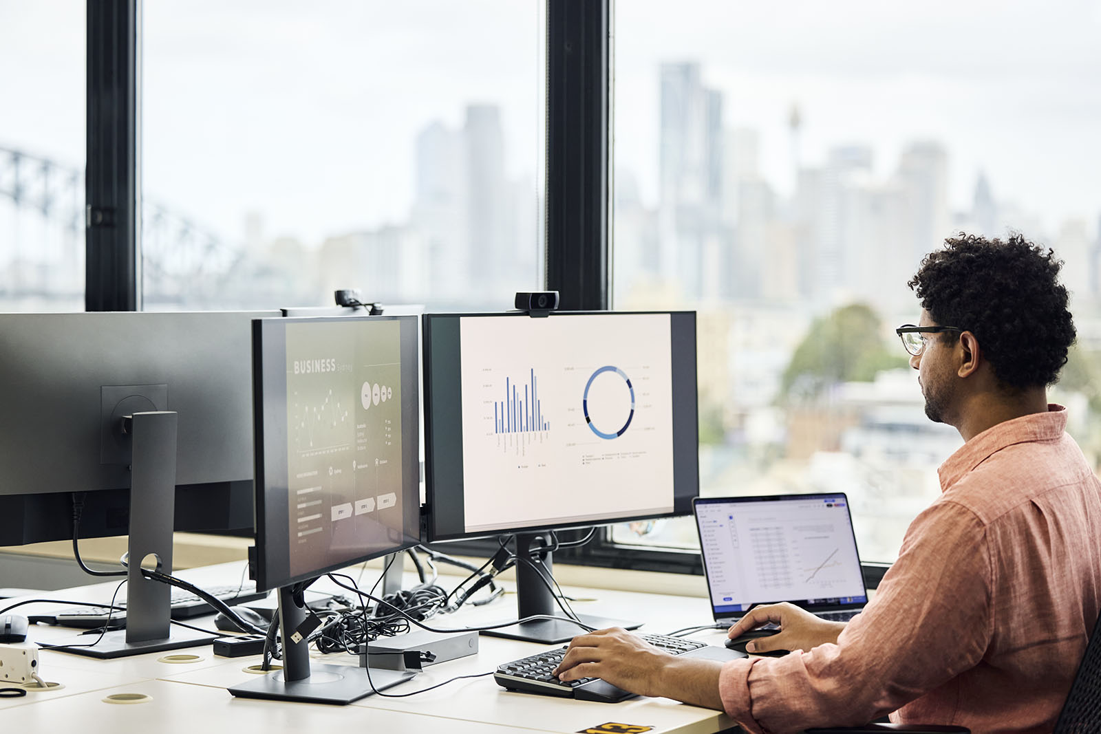 A professional sitting at a desk in a modern office, working on a laptop connected to dual monitors displaying business analytics dashboards, with large windows revealing a blurred city skyline and bridge in the background.