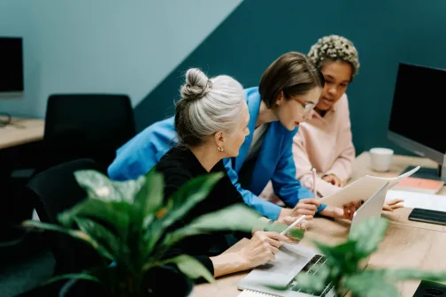Three coworkers gathered around a desk reviewing documents together, collaborating in a modern office setting.
